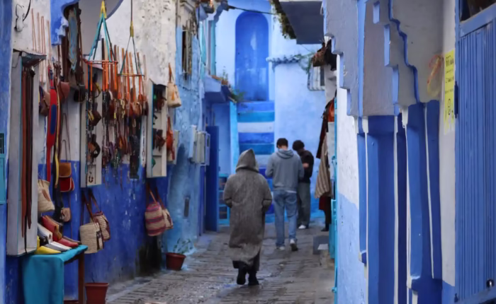 The blue streets of Chefchaouen with people walking in the street. the image shows also 3 men one of them is wearing th Moroccan long dress Jelaba. Moroccan Travel - Morocco travel packages - Morocco holidays - Morocco vacations package - tour in Morocco - trips to morocco / viaggio in morocco