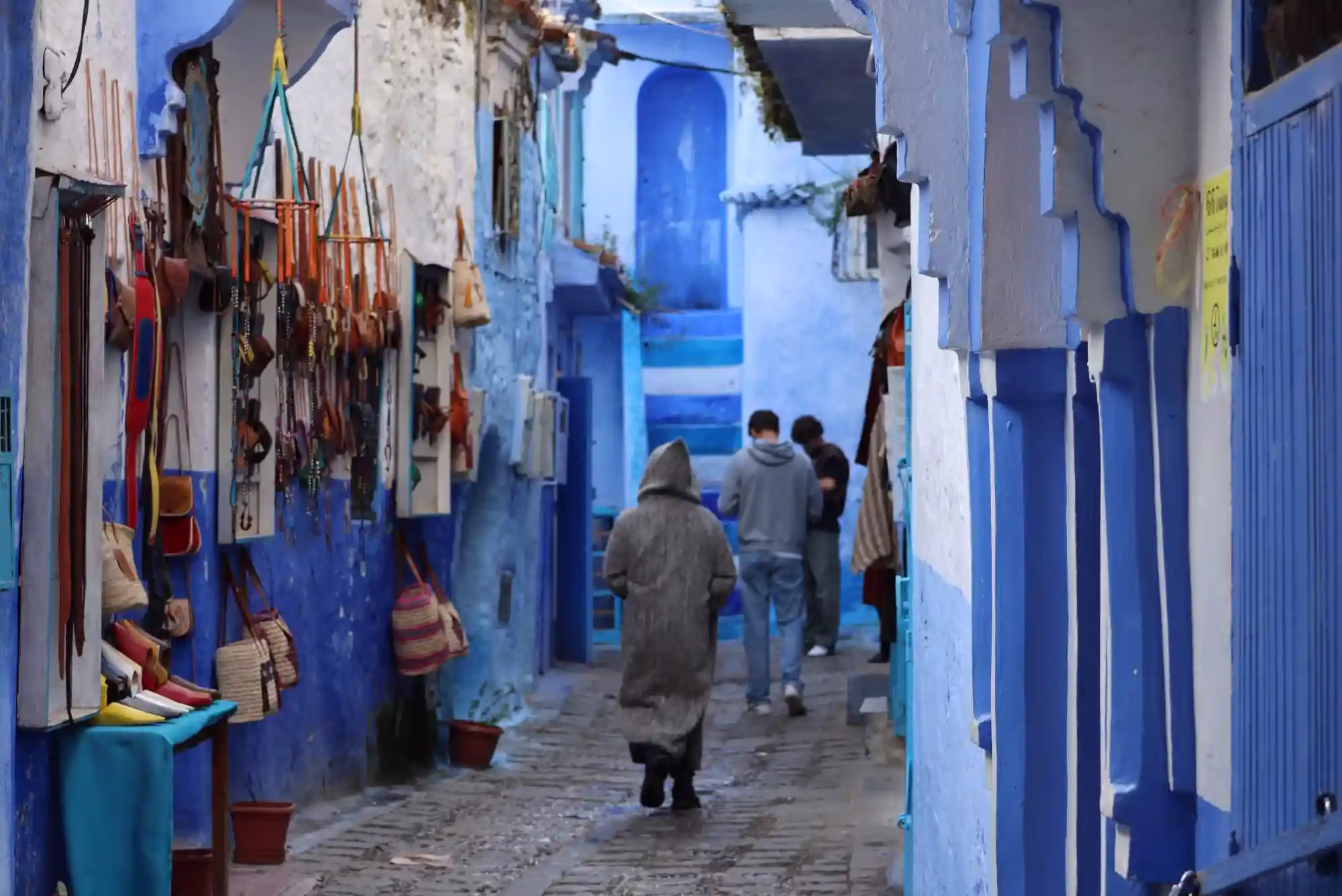 The blue streets of Chefchaouen with people walking in the street. the image shows also 3 men one of them is wearing th Moroccan long dress Jelaba. Moroccan Travel - Morocco travel packages - Morocco holidays - Morocco vacations package - tour in Morocco - trips to morocco / viaggio in morocco
