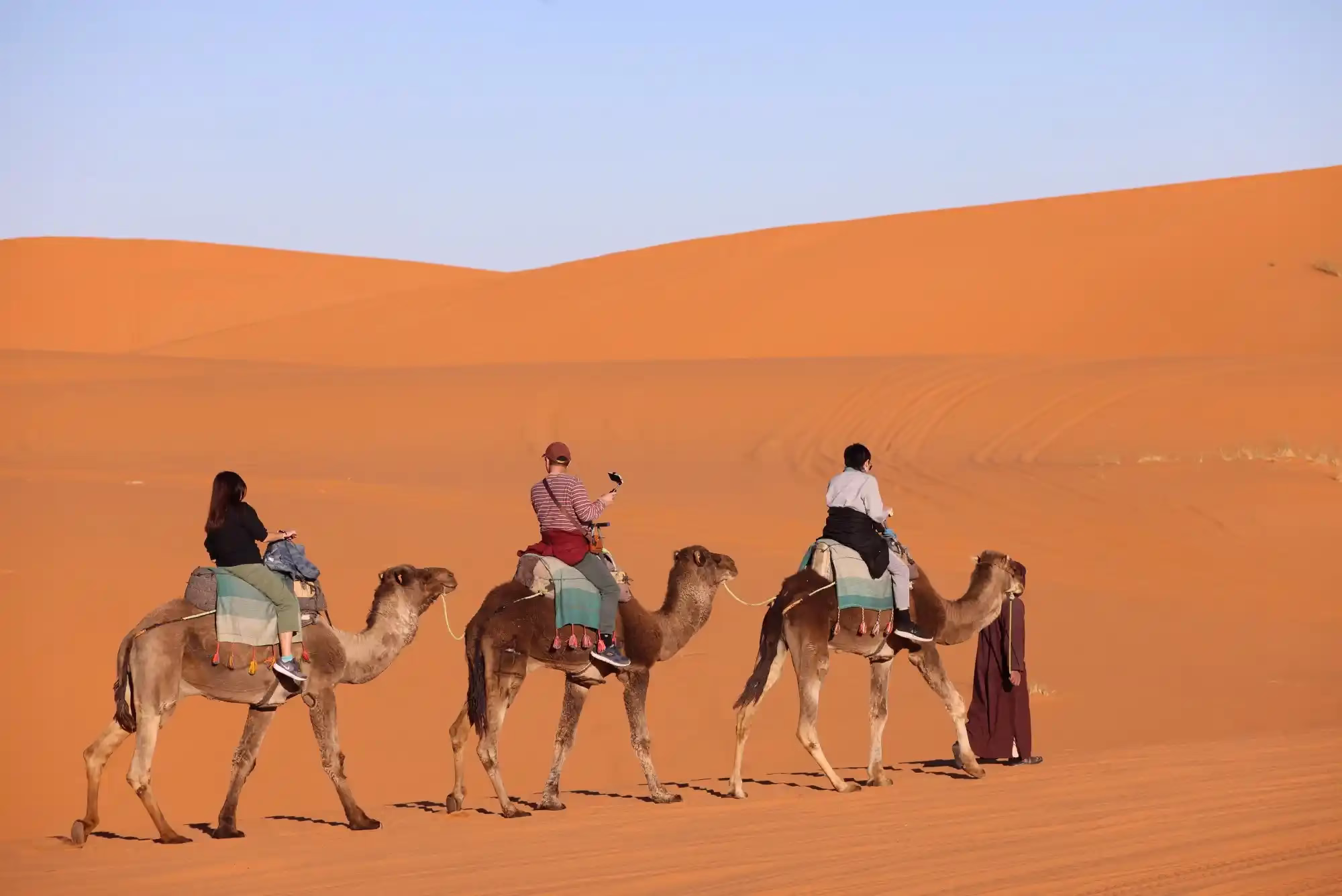 3 clients on a camel ride with guide in the middle of the golden dunes of Sahara Desert (Erg chebbi) during our 5 day Morocco tour from Casablanca to Marrakech - Morocco tour package - Morocco holiday - Morocco travel package - Morocco vacation package - Viaggio in Marocco - 5 giorni da Casablanca - Moroccan Travel packages .